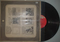 Vinyl record with a red label and a brown paper sleeve on a gray wall.